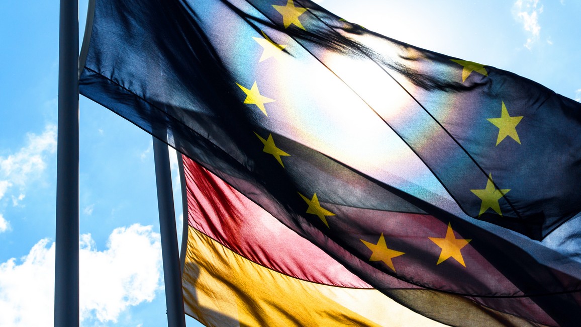 Eine EU-Flagge flattert im Wind und verdeckt leicht eine Deutschland-Flagge, die dahintersteht. Im Hintergrund scheint die Sonne vor einem hellblauen, leicht bewölkten Himmel.