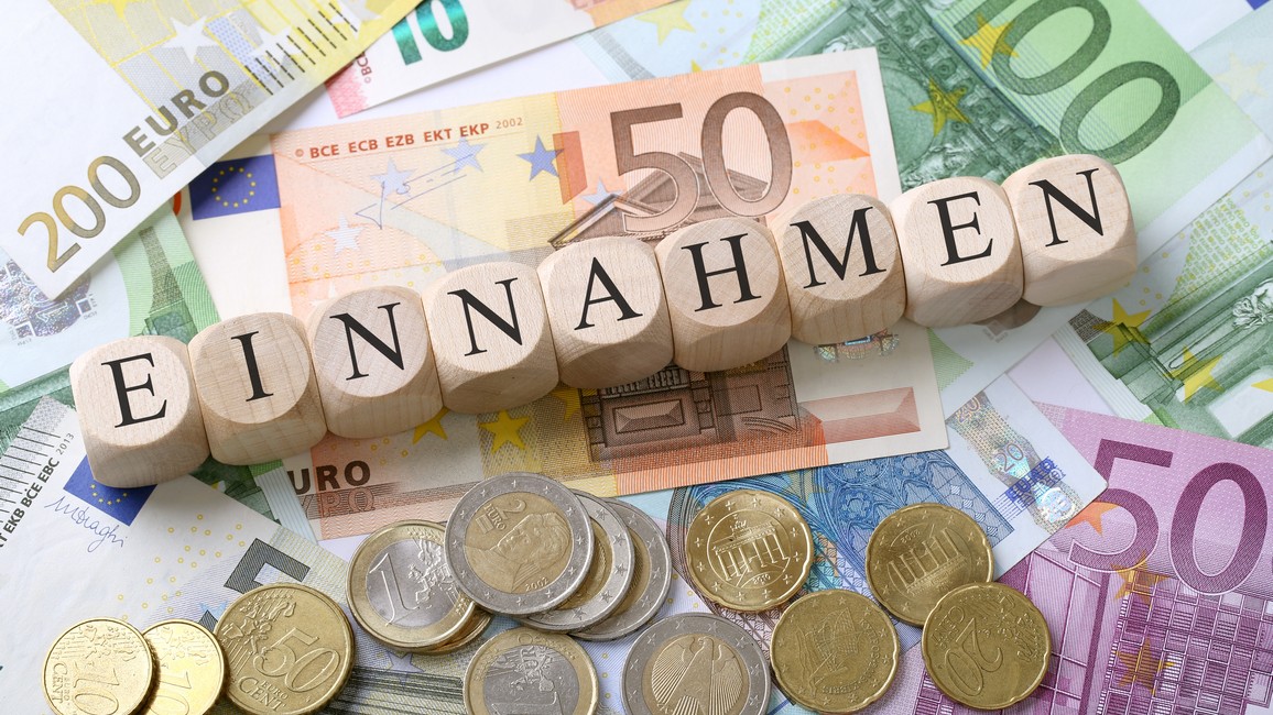 Holzwürfel, die zusammen das Wort „Einnahmen“ bilden, liegen gemeinsam mit einigen Euro-Münzen auf Euro-Geldscheinen.
