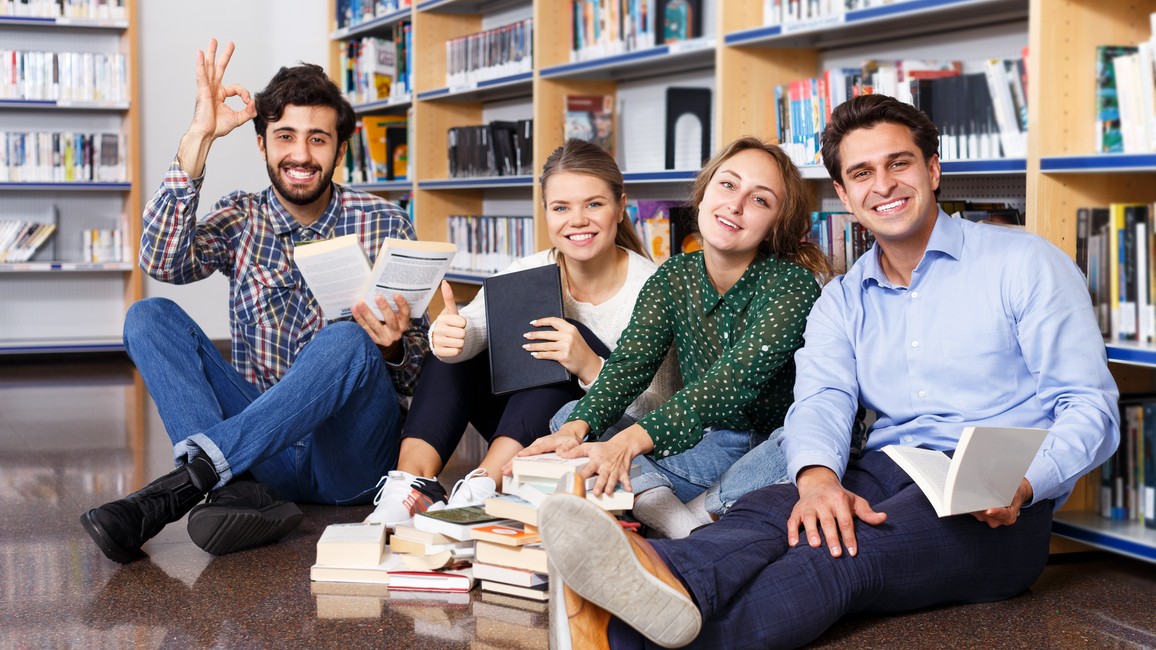 Fröhliche junge Menschen verschiedener Ethnien sitzen auf dem Fußboden in einer Bibliothek, umgeben von Büchern.