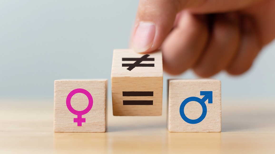 Drei Holzwürfel sind nebeneinander auf einem Tisch angeordnet. Der linke zeigt das Symbol für Frauen, der rechte das für Männer. In der Mitte wird von einer Hand das Ungleich-Zeichen in ein Gleich-Zeichen umgekippt.