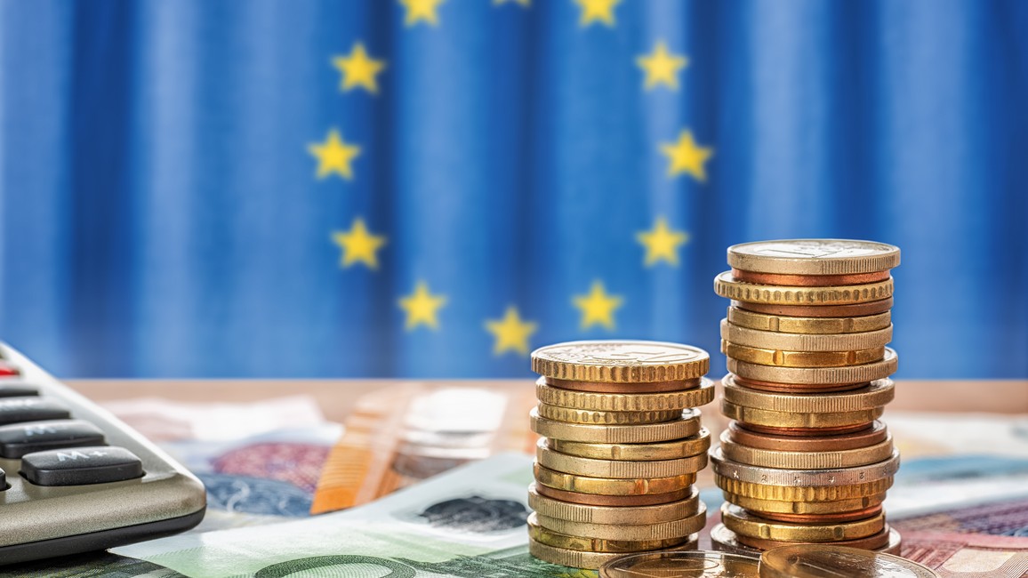 Vor einem verschwommenen blauen Hintergrund mit EU-Sternen sind auf einem Tisch Münzen und Euro-Geldscheine zu sehen sowie am linken Bildrand ausschnittsweise ein Tischrechner.