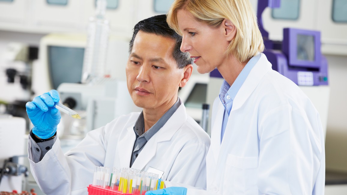 Zwei Wissenschaftler im Laborkittel – ein Mann und eine Frau – betrachten gemeinsam ein schmales Reagenzglas mit gelber Flüssigkeit. Die Frau hält einen mit vielen Reagenzgläsern gefüllten Ständer vor sich. Im Hintergrund sind medizintechnische Geräte zu sehen.